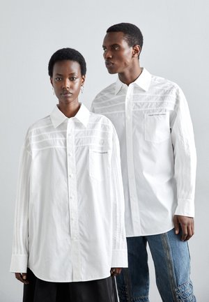 Hvite oversized skjorter med knapper har vertikale stripe detaljer og sidelommer, og de er kombinert med kontrasterende blå denimbukser.