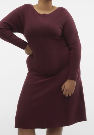 Persona con un vestido burdeos ajustado, de manga larga y largo hasta la rodilla, de pie con una mano en la cadera y la otra relajada.