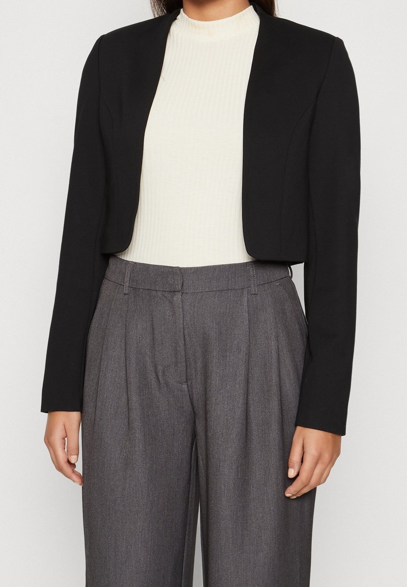 Femme portant un blazer court noir, un haut côtelé blanc et un pantalon gris taille haute à plis, debout devant un fond uni.
