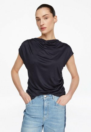 Donna con capelli raccolti che indossa una maglia blu navy a maniche corte drappeggiata e jeans azzurri a vita alta, con le mani nelle tasche.