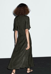 Dunkelolivgrünes Kleid mit kurzen Ärmeln, fließender Silhouette und strukturiertem Stoff. Lange Länge mit Seitenschlitzen, kombiniert mit Sandalen.