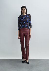 Haut noir à manches longues avec un motif floral bleu, associé à un pantalon ajusté bordeaux, complété par des chaussures plates noires sur un fond neutre.