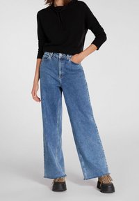 Breda blå jeans i denim, med hög midja och rå kant. Kombineras med en svart hoodie och bruna platåskor.