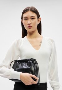 Νεαρό άτομο με μακριά σκούρα μαλλιά κρατάει μαύρο δερμάτινο τσαντάκι με κορδόνι, φορώντας λευκό μακρυμάνικο μπλουζάκι με διαφανείς μανίκια και μαύρο παντελόνι.