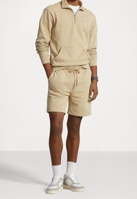 Tan fleece-tröja med kvartsdragkedja och känguruficka, tillsammans med matchande shorts. Vita sneakers med beige detaljer och texturerade strumpor.