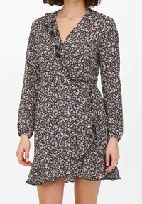 Žena v černých dlouhých rukávech oblečená v zavinovacích šatech s malým růžovým a bílým květinovým vzorem a volánkovými detaily.