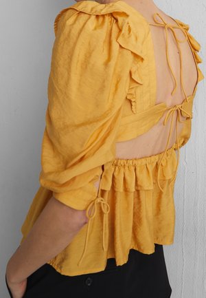 Gele blouse met ruches, volumineuze mouwen en een open achterkant met meerdere strikken, gedragen met zwarte broek tegen een neutrale achtergrond.