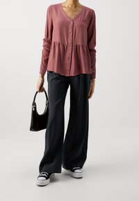 Top a maniche lunghe color rosa con scollatura a V, bottoni sul davanti e vita a peplo. Abbinato a pantaloni larghi neri e sneakers.