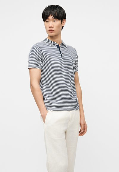 Homme portant un polo à manches courtes rayé bleu marine et blanc avec un pantalon blanc, debout, une main dans la poche, regardant vers la gauche.