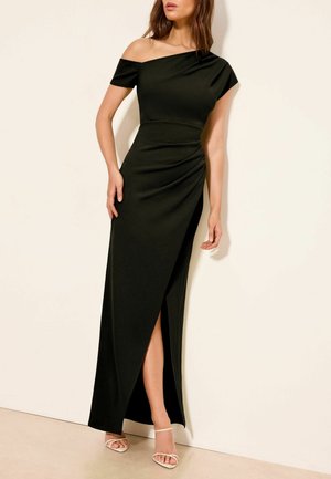 Kvinde iført en sort, asymmetrisk gulvlængde kjole med høj slids og hvide, strop-sandaler foran en ensfarvet væg.