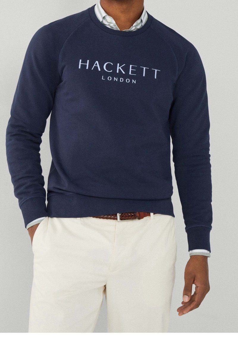 Homme portant un sweat-shirt Hackett London bleu marine sur une chemise blanche avec un pantalon beige et une ceinture tressée marron.