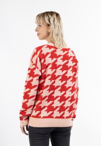 Svetr v červeném a růžovém vzoru houndstooth s dlouhými rukávy, měkkou texturou a žebrovanými manžetami, navržený v uvolněném střihu.