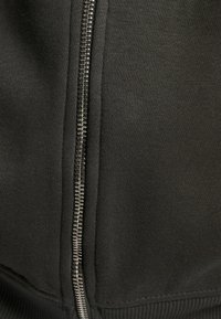 Musta vetoketjullinen huppari, jossa on sileä pinta, metallivetoketju, ribatoi hihansuut ja helma. Kangas on paksu ja pehmeä.