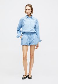 Γυναίκα που στέκεται, φοράει μπλε ριγωτό πουκάμισο με μακριά μανίκια και ψηλόμεσα σορτς που ταιριάζουν, με μαύρα παπούτσια Mary Jane σε λευκό φόντο.