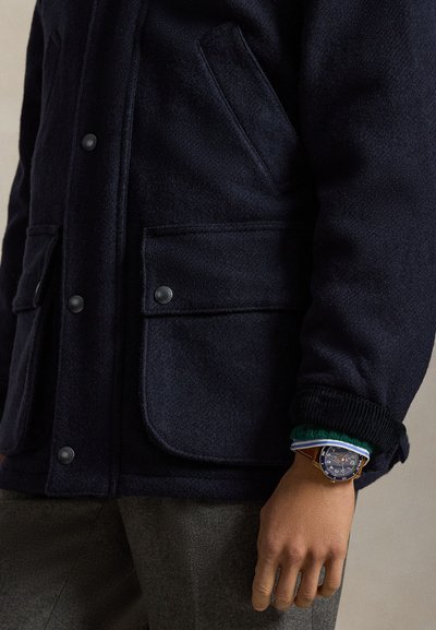 Mörkblå ullrock med två framfickor och tryckknappar, kombinerad med en månglagrad manschett och en brun läderarmbandsur.