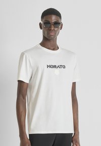 Camiseta de algodón blanca con la palabra "MORATO" en diseño negro y rayas, con un logo circular debajo. Mangas cortas, cuello redondo.