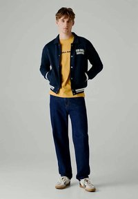 Mørkeblå varsity-jakke med hvite striper, logo på brystet, gul t-skjorte under, mørke jeans, og hvite sneakers med gummisåler.