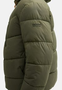Oliwkowa kurtka puchowa z pikowanymi sekcjami, zamkniętą kieszenią i logo. Wykonana z gładkiego, lekkiego materiału.
