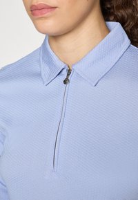 Camisa azul claro com fecho éclair, feita de tecido texturizado, apresentando um pequeno fecho de metal prateado e um design de colarinho estruturado.