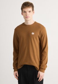 Μακρυμάνικη καφέ βαμβακερή πουκάμισο με υφασμάτινο σχέδιο, ριμπ μανσέτες και μικρή λογότυπη επιφάνεια στο στήθος. Τυπικός σχεδιασμός με στρογγυλή λαιμόκοψη.