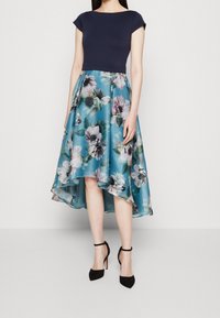Marinblå överdel med kort ärm, blommönstrad kjol i teal och vitt, asymmetrisk fåll, i kombination med svarta högklackade ankelskor med rem.