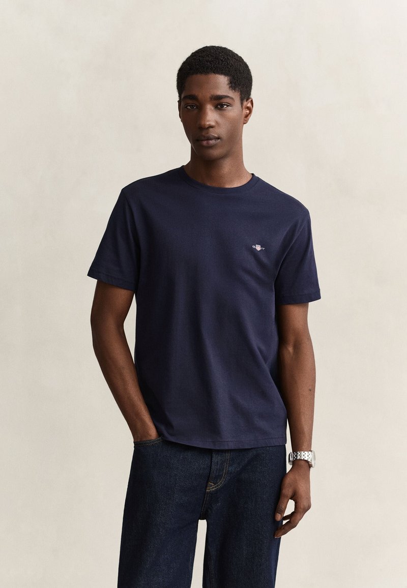 Giovane uomo che indossa una maglietta blu navy con piccolo logo sul petto, jeans scuri e un orologio da polso argentato, in piedi davanti a uno sfondo chiaro uniforme.