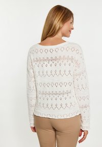 Maglione bianco a maglia con pizzo, caratterizzato da un motivo geometrico e un colletto rotondo. Maniche lunghe e design leggermente accorciato. Indossato sopra pantaloni beige.