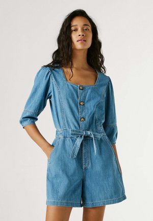 Donna che indossa un romper di jeans blu con bottoni, maniche a 3/4 e vita annodata, in piedi con le mani nelle tasche davanti a uno sfondo semplice.