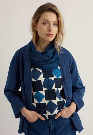 Donna con capelli biondi che indossa una sciarpa blu, una maglia con stampa a quadri nera e blu, una giacca blu e jeans blu, con le mani nelle tasche.