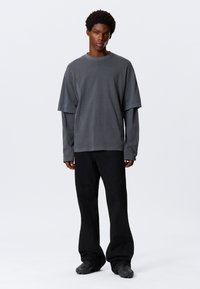 Szara, przewiewna koszulka noszona na długim rękawie, połączona z czarnymi spodniami o szerokich nogawkach i fakturowanymi czarnymi butami. Gładkie tło.