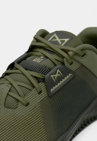 Πράσινο αθλητικό παπούτσι με υφή στην επάνω επιφάνεια, επίπεδα κορδόνια και πλαστική προστασία κορδονιών. Ο σχεδιασμός περιλαμβάνει διακριτά οριζόντια και κάθετα σχέδια.