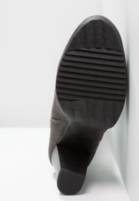 Bota de tacón alto de ante gris con suela de goma negra texturizada y prominentes surcos, que presenta un diseño elegante y una punta redondeada.