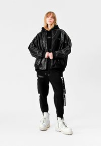 Chaqueta de cuero negra de gran tamaño con bolsillos con cremallera, combinada con pantalones negros que tienen cremalleras laterales y botas blancas con cordones para un contraste.