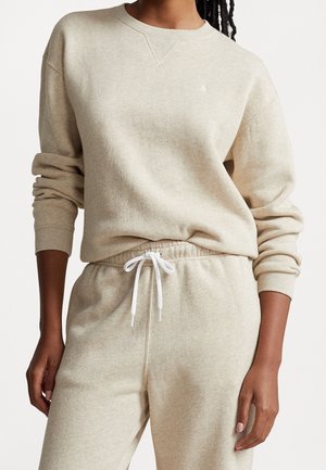 Personne portant un sweat-shirt beige à manches longues et un pantalon de jogging assorti avec un cordon blanc, debout avec un bras relâché le long du corps.