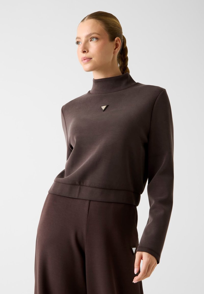 Vrouw met gevlochten haar draagt een donkerbruine hoge nek top met lange mouwen en bijpassende broek met kleine driehoekige logo's.