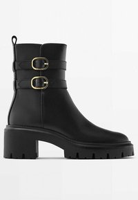 Stivaletto in pelle nera con due fibbie dorate, tacco spesso e suola testurizzata. Presenta una finitura liscia e un inserto sul retro.