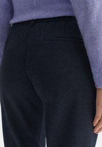 Detailní záběr na osobu v tmavých strukturovaných kalhotách a světle fialovém pleteném svetr, s viditelnou zadní kapsou a uvolněnou rukou.