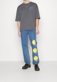 Camiseta gris de gran tamaño con logo, vaqueros azules con tres grandes impresiones de cara sonriente amarilla, y zapatos negros con cordones.