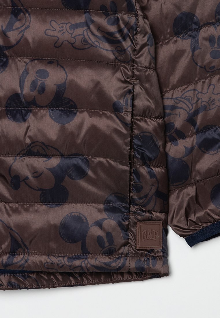 Chaqueta acolchada marrón con un patrón repetido de Mickey Mouse, diseño acolchado y un parche del logo de GAP en el dobladillo. Acentos en azul marino en los puños.