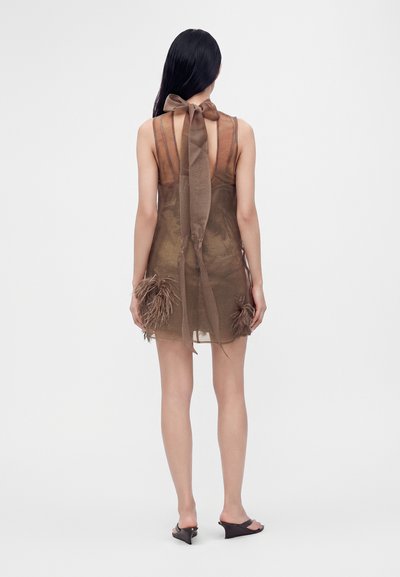 Femme aux longs cheveux noirs portant une robe marron transparente sans manches avec un nœud au cou et des accents de plumes, debout pieds nus sur un fond clair.