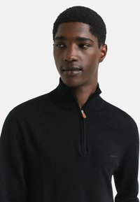 Zwarte zip-up trui van zacht materiaal, met een hoge kraag, een klein logo op de borst en zilverkleurige hardware op de rits.