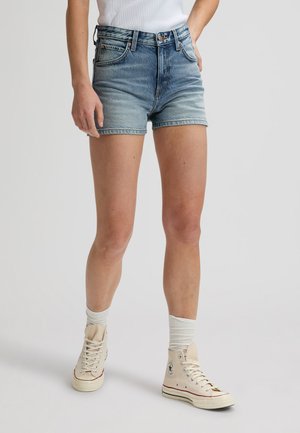 Személy, aki kék farmer rövidnadrágot, fehér zoknit és bézs magas szárú tornacipőt visel, egy egyszínű világos háttér előtt áll.