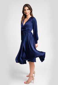 Granatowa satynowa sukienka z głębokim dekoltem w kształcie V, długimi rękawami i związanym w pasie. Falująca spódnica podkreśla ruch. Noszona z cielistymi sandałami na obcasie.