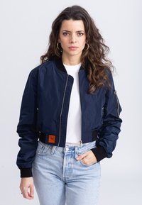 Giacca bombardiere navy con polsini a costine, zip frontale e etichetta arancione sull'orlo. Indossata sopra una maglietta bianca e abbinata a jeans azzurri chiari.