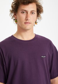 Paarse katoenen t-shirt met een ronde hals, voorzien van een klein zwart-wit logo op de linkerborst. Zachte textuur en een ontspannen pasvorm.