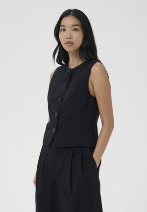 Kobieta o długich czarnych włosach, ubrana w czarną bezrękawnikową kamizelkę zapinaną na guziki i czarne spodnie, stojąca z jedną ręką w kieszeni na szarym tle.