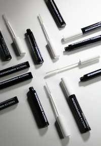 Black and white lash serum tubes with distinct branding. Tubes are cylindrical; some have white caps. Brushes are straight and slender.