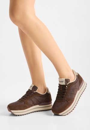 Gambe che indossano scarpe da ginnastica Woden marroni con accenti beige, lati trapuntati, suola spessa a righe bianche e lacci neri su sfondo bianco uniforme.