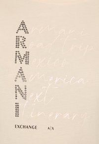 Ανοιχτό �μπεζ μπλουζάκι με μεταλλικά τρουκς που σχηματίζουν τη λέξη "ARMANI" κάθετα, με καλλιγραφικά λόγια "Road Trip Mexico America Next Itinerary" από κάτω.