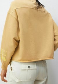 Persona con un sudadera beige suelta y corta con texto amarillo en la manga y pantalones blanco rotos con un gran bolsillo trasero con botones.